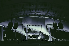 A large stage setup featuring an array of lighting equipment and large hanging speakers under an expansive canopy. The structure includes metal beams and pillars, illuminated with various lights focusing on the stage area.