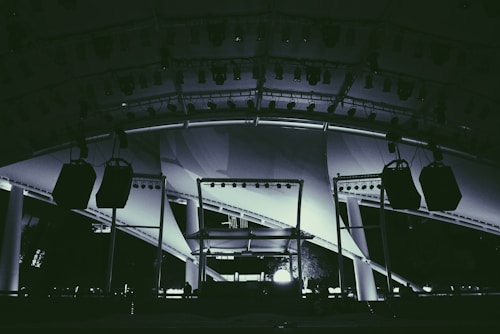 A large stage setup featuring an array of lighting equipment and large hanging speakers under an expansive canopy. The structure includes metal beams and pillars, illuminated with various lights focusing on the stage area.