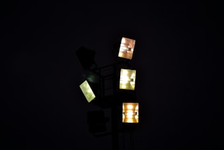 A cluster of illuminated floodlights against a dark background, forming geometric patterns with their square shapes. The lights emit a range of colors from yellow to white.