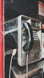 An old, dusty public payphone with cobwebs covering its corners. It has a metal body with a black handset hanging on a hook. The surrounding area shows signs of neglect, with a red frame and dark background.