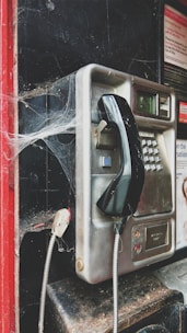 An old, dusty public payphone with cobwebs covering its corners. It has a metal body with a black handset hanging on a hook. The surrounding area shows signs of neglect, with a red frame and dark background.