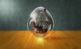 A transparent glass orb rests on a wooden surface, containing a swirling, dark substance with intricate patterns. The orb reflects light, causing a glow on the surface beneath it. The background is a soft, blurred gradient, emphasizing the orb as the focal point.