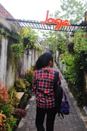 A person wearing a red plaid shirt and carrying a colorful backpack walks down a narrow stone pathway surrounded by lush greenery and hanging plants. Above the pathway, there is a structure with a sign that says 'Jogja' in bright red and yellow letters.
