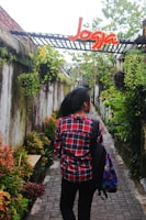 A person wearing a red plaid shirt and carrying a colorful backpack walks down a narrow stone pathway surrounded by lush greenery and hanging plants. Above the pathway, there is a structure with a sign that says 'Jogja' in bright red and yellow letters.
