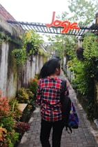 A person wearing a red plaid shirt and carrying a colorful backpack walks down a narrow stone pathway surrounded by lush greenery and hanging plants. Above the pathway, there is a structure with a sign that says 'Jogja' in bright red and yellow letters.