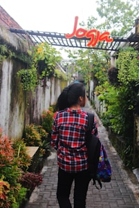 A person wearing a red plaid shirt and carrying a colorful backpack walks down a narrow stone pathway surrounded by lush greenery and hanging plants. Above the pathway, there is a structure with a sign that says 'Jogja' in bright red and yellow letters.