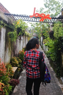 A person wearing a red plaid shirt and carrying a colorful backpack walks down a narrow stone pathway surrounded by lush greenery and hanging plants. Above the pathway, there is a structure with a sign that says 'Jogja' in bright red and yellow letters.