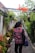 A person wearing a red plaid shirt and carrying a colorful backpack walks down a narrow stone pathway surrounded by lush greenery and hanging plants. Above the pathway, there is a structure with a sign that says 'Jogja' in bright red and yellow letters.