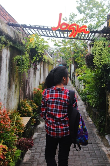 A person wearing a red plaid shirt and carrying a colorful backpack walks down a narrow stone pathway surrounded by lush greenery and hanging plants. Above the pathway, there is a structure with a sign that says 'Jogja' in bright red and yellow letters.
