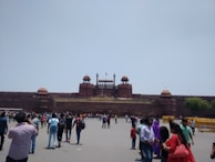A large historical red sandstone fort with distinctive architecture, featuring multiple domes and a central flag. Crowds of people are gathered in front of the structure, engaging in various activities and walking around.