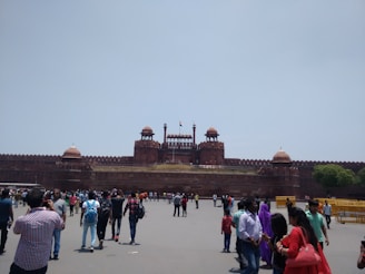 A large historical red sandstone fort with distinctive architecture, featuring multiple domes and a central flag. Crowds of people are gathered in front of the structure, engaging in various activities and walking around.