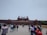 A large historical red sandstone fort with distinctive architecture, featuring multiple domes and a central flag. Crowds of people are gathered in front of the structure, engaging in various activities and walking around.