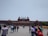 A large historical red sandstone fort with distinctive architecture, featuring multiple domes and a central flag. Crowds of people are gathered in front of the structure, engaging in various activities and walking around.