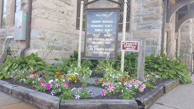 A wooden signboard displaying church service times is nestled in a small garden bed. The garden features vibrant flowers in shades of pink, purple, white, and orange, surrounded by green foliage. Nearby, a smaller sign indicates the church office. The stone exterior of a building serves as the backdrop.