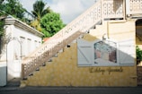 A yellow wall with decorative white shutters flanks a staircase with ornate railing designs. A painted mural is framed between the shutters, depicting a pastoral scene. Lush green foliage and palm trees are visible in the background, casting shadows on the architecture. The name 'L'Auberge Gourmande' is scripted on the wall.