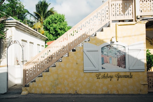 A yellow wall with decorative white shutters flanks a staircase with ornate railing designs. A painted mural is framed between the shutters, depicting a pastoral scene. Lush green foliage and palm trees are visible in the background, casting shadows on the architecture. The name 'L'Auberge Gourmande' is scripted on the wall.