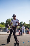 A person rollerblades skillfully on a path, maneuvering between small blue cones. They wear a striped shirt with suspenders and a cap. In the background, families and children with strollers enjoy the sunny day in an outdoor setting.