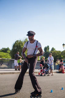 A person rollerblades skillfully on a path, maneuvering between small blue cones. They wear a striped shirt with suspenders and a cap. In the background, families and children with strollers enjoy the sunny day in an outdoor setting.