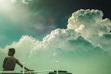 A person stands on a platform or balcony, gazing at large, dramatic clouds that fill the sky. The sun is shining brightly on the left, casting a soft glow. The scene conveys a sense of tranquility and contemplation.