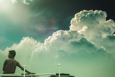 A person stands on a platform or balcony, gazing at large, dramatic clouds that fill the sky. The sun is shining brightly on the left, casting a soft glow. The scene conveys a sense of tranquility and contemplation.