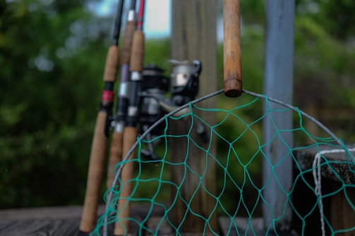 Several fishing rods are standing upright, leaning against a wooden post with their handles facing outward. In the foreground, a green fishing net with a wooden handle is visible, slightly out of focus. The background consists of blurred greenery, suggesting an outdoor setting.