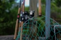 Several fishing rods are standing upright, leaning against a wooden post with their handles facing outward. In the foreground, a green fishing net with a wooden handle is visible, slightly out of focus. The background consists of blurred greenery, suggesting an outdoor setting.