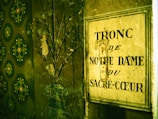 A wall with textured wallpaper featuring green floral patterns is partially visible. Next to it is an old metallic sign with the French words 'Tronc de Notre Dame du Sacré-Coeur' painted on it, indicative of a donation box. The sign and wall appear worn and aged, providing a vintage and historical feel.