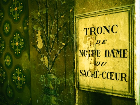 A wall with textured wallpaper featuring green floral patterns is partially visible. Next to it is an old metallic sign with the French words 'Tronc de Notre Dame du Sacré-Coeur' painted on it, indicative of a donation box. The sign and wall appear worn and aged, providing a vintage and historical feel.