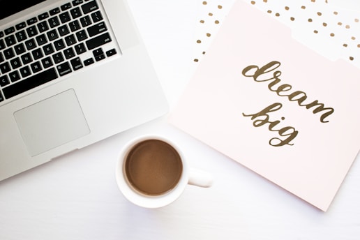 A workspace setting with a laptop, a cup of coffee, and a folder with the phrase 'dream big' written on it. The background features a light color palette with subtle decorative dots.