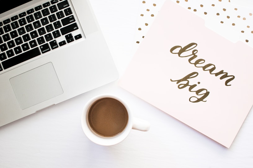 A workspace setting with a laptop, a cup of coffee, and a folder with the phrase 'dream big' written on it. The background features a light color palette with subtle decorative dots.