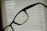 A close-up of a pair of eyeglasses with water droplets on the lenses, set against a blurred background of blinds or a window with a soft light filtering through.