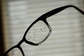A close-up of a pair of eyeglasses with water droplets on the lenses, set against a blurred background of blinds or a window with a soft light filtering through.