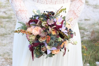 A person wearing a lace-sleeved white dress holding an elaborate bouquet of flowers. The bouquet contains a mix of roses, wildflowers, and various green foliage with colors ranging from peach and lavender to deep red.