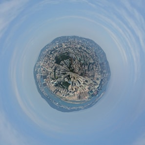 A tiny planet effect transforms a panoramic cityscape into a spherical view. The densely packed buildings and skyscrapers encircle a central point, creating a miniature world of urban architecture. A section of water and hills surrounds the city, adding natural elements to the scene.
