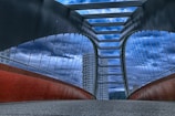 A modern bridge with tall, arched supports and a series of horizontal beams spans across, converging towards the horizon. The structure is framed by tall, glass-covered buildings in the background, under a dramatic, cloudy sky. The underside of the bridge features a red, curving element, while the pathway is covered with a textured surface.