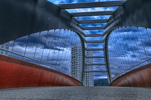 A modern bridge with tall, arched supports and a series of horizontal beams spans across, converging towards the horizon. The structure is framed by tall, glass-covered buildings in the background, under a dramatic, cloudy sky. The underside of the bridge features a red, curving element, while the pathway is covered with a textured surface.