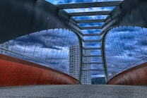 A modern bridge with tall, arched supports and a series of horizontal beams spans across, converging towards the horizon. The structure is framed by tall, glass-covered buildings in the background, under a dramatic, cloudy sky. The underside of the bridge features a red, curving element, while the pathway is covered with a textured surface.