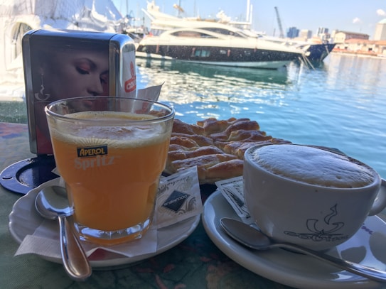 A glass of orange Aperol Spritz and a cup of frothy cappuccino sit on a table by the waterfront. A slice of pastry is placed between them. The background features a marina with several luxury yachts docked, and the reflection of the blue sky and buildings on the water is visible.