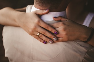 Two hands with manicured nails are gently placed on the back of a person wearing a light-colored suit jacket. One hand has a gold wedding ring, and the nails are painted in dark purple and glittery silver colors.