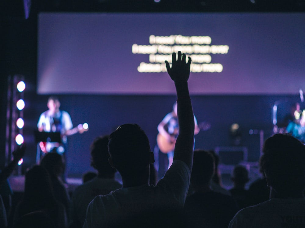 man wearing white t-shirt raising right hand on moshpit while concert is going on inside dim lit venue