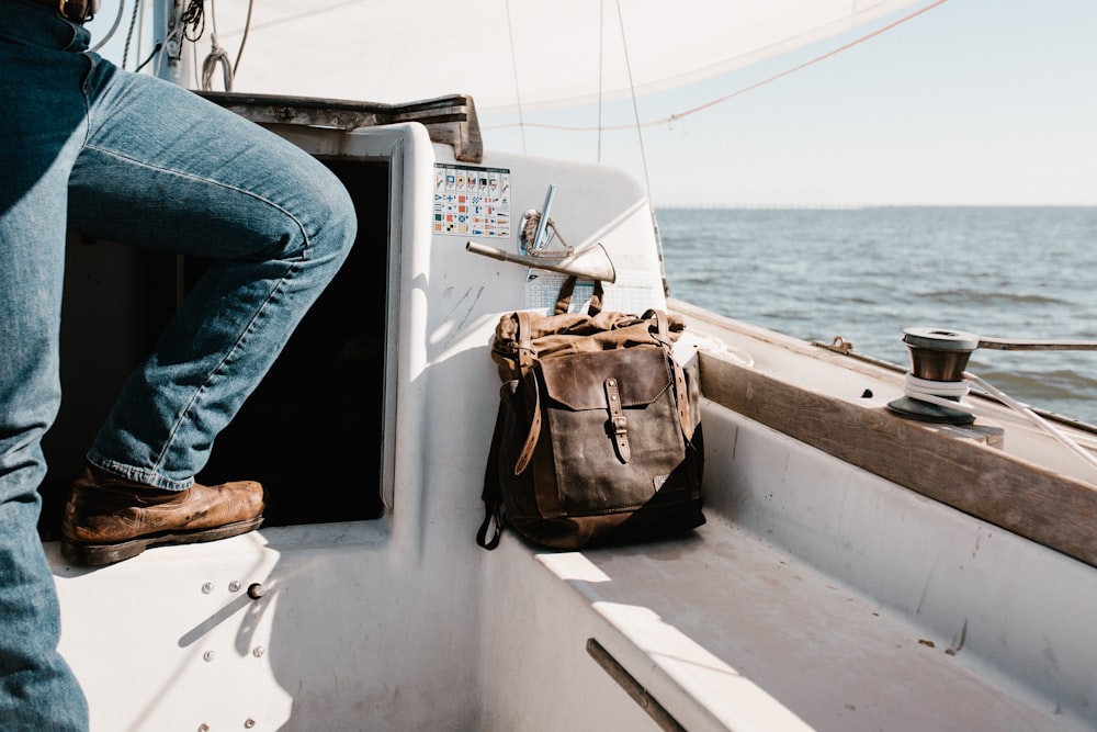 Person in blauer Jeans auf Yacht neben brauner Tasche