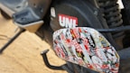 A close-up view of a motorcycle or scooter's footrest area, exhibiting a collage of various colorful stickers. The stickers include text and diverse designs, contributing a vibrant look. The background includes part of the vehicle with a visible red UNI logo and other mechanical components.