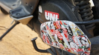 A close-up view of a motorcycle or scooter's footrest area, exhibiting a collage of various colorful stickers. The stickers include text and diverse designs, contributing a vibrant look. The background includes part of the vehicle with a visible red UNI logo and other mechanical components.