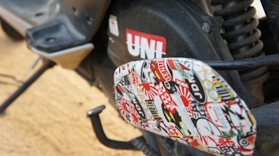 A close-up view of a motorcycle or scooter's footrest area, exhibiting a collage of various colorful stickers. The stickers include text and diverse designs, contributing a vibrant look. The background includes part of the vehicle with a visible red UNI logo and other mechanical components.