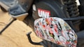 A close-up view of a motorcycle or scooter's footrest area, exhibiting a collage of various colorful stickers. The stickers include text and diverse designs, contributing a vibrant look. The background includes part of the vehicle with a visible red UNI logo and other mechanical components.
