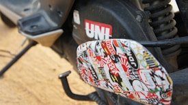 A close-up view of a motorcycle or scooter's footrest area, exhibiting a collage of various colorful stickers. The stickers include text and diverse designs, contributing a vibrant look. The background includes part of the vehicle with a visible red UNI logo and other mechanical components.