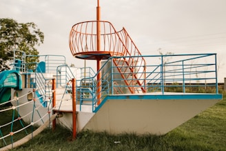 A playground structure designed like a ship with blue and orange railings on a grassy field. It features stairs, rope ladders, and climbing areas, surrounded by open space and a few trees in the background.