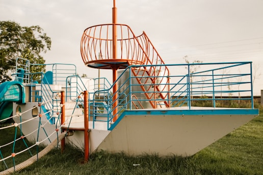 A playground structure designed like a ship with blue and orange railings on a grassy field. It features stairs, rope ladders, and climbing areas, surrounded by open space and a few trees in the background.