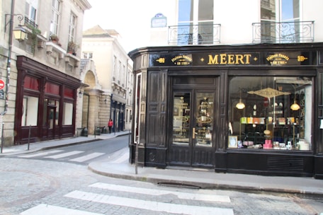 A corner building with a storefront displaying the name 'Meert' in elegant lettering. The shop facade is black with large windows showcasing various products. A narrow street curves alongside, bordered by historic stone buildings with decorative windows and balconies. A pedestrian and a cyclist can be seen down the street, adding a sense of quiet activity.