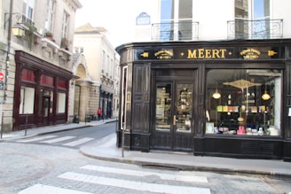 A corner building with a storefront displaying the name 'Meert' in elegant lettering. The shop facade is black with large windows showcasing various products. A narrow street curves alongside, bordered by historic stone buildings with decorative windows and balconies. A pedestrian and a cyclist can be seen down the street, adding a sense of quiet activity.